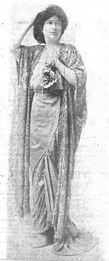 A white woman standing, one hand on her head, one hand holding flowers on her chest, wearing a flowing gown and shawl