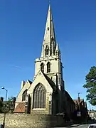 A handsome stone church showing a wide window beyond which is a large tower with a spire and a single crocketted pinnacle