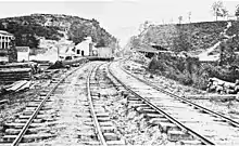 Black and white photo shows railroad tracks cutting through a gap between two high hills.