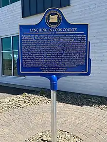 Large historical marker, blue with gold lettering. First line says, "Lynching in Coos County". An emblem says "Community Remembrance Project" at the top.