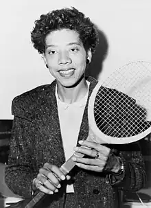 A woman looking toward the camera with a tennis racket held diagonally in front of her.