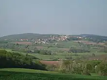 A small village with a visible church tower, on a low hill surrounded by fields and trees, seen from another such hill