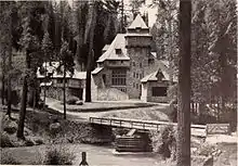 A monochrome photograph of a 75-foot-tall building with steeply pitched roof elements, seen amid tall pine trees from across a river. A rustic one-lane bridge over the river is in the foreground.