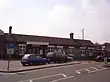 A long building with two chimneys protruding from its roof and four cars driving from left to right across a paved area in the foreground