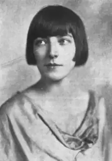 A young white woman with bobbed hair cut with bangs, wearing a satin top with a scooped neckline