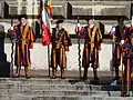 Picture of the Pontificial Swiss Guard. Three guards are in the center of the picture wearing traditional clothing with orange and blue stripes. They also wear black berets. The middle guard holds a pole with the flag of the guard. The two other guards flank him on both sides. They hold two long two-handed swords in their right hand pointed to the ground. The blades of the swords are waved, shaped in a zigzag manner with rounded edges.