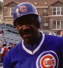 A man, wearing a baseball cap with the Chicago Cubs "red C" logo in the center and a blue baseball uniform with the Cubs logo to his left, faces forward.