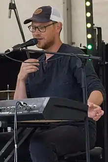 Bespectacled person wearing a hat while sitting at a keyboard and speaking into a microphone