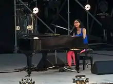 A woman with black glasses seated at a grand piano on a stage, wearing a blue singlet, pink tights and sneakers.