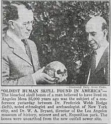 Black-and-white newspaper photo of two scientists looking down at an ancient human skull