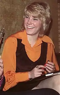 A young woman with blonde hair, wearing an orange blouse and a black vest