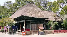 Three-quarter view of a wooden building with a thatched hip roof. Another structure of similar style extends from the back of the building.