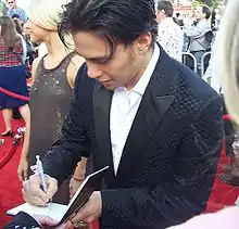 A man in a tuxedo is looking down as he signs an autograph on the red carpet. There are people standing in the background.