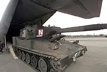 An AGS in woodland camouflage pattern rolls out of the rear cargo ramp of a C-130. The gun is forward. There is no armament mounted on the commander's hatch.