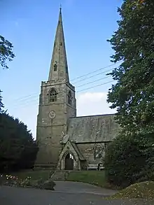 A tall spire is on the left and part of the body of the church is on the right. Steps lead to a porch and the whole is framed by trees