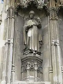 Stone statue of a standing man wearing robes and a mitre, carrying a tablet in one hand and holding his other hand up.