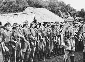 Soldiers on parade in front of a hut in a tropical setting. An officer in a steel helmet with a walking stick stands in front facing away from them, while the men behind him are wearing a various assortment of uniforms including steel helmets, slouch hats, shorts and are carrying rifles