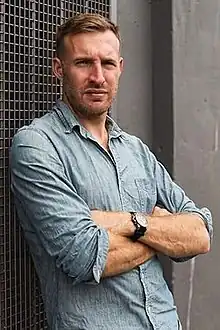 Photo of author Owen Laukkanen in a pale blue denim shirt leaning up against an industrial building in Vancouver, BC.  His arms are crossed over his chest and his shirt sleeves are rolled up revealing a black watch. His brow is furrowed and he has a serious look on his face. His body is slightly angled away from the camera. His hair is light brown and short and he has short, neat facial hair.