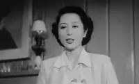 An attractive young Japanese woman, wearing a white blouse, is shown talking, photographed from below; a lamp, some bottles on a mantlepiece and part of a painting are visible in the left background.