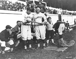 Two men smiling and holding a baseball bat, with a child next to them. Several seated men are sitting behind them, in front of grandstands.