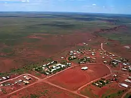  an aerial view of a small settlement, showing about seventy rooftops, red dirt roads and a dirt oval, with dry scrubland receding into the distance