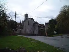 Stone building with slit windows and battlements. Foreground is road with grass verges.