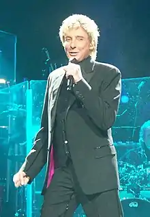 A blond-haired man wearing a dark suit, singing into a microphone