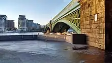 View of an empty paved area that is part of a public riverside footpath, with a railway bridge, next, to the right.  Backdrop of the river Thames.