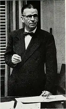 Mervin Kelly, wearing a dark suit and bow tie, standing behind a desk