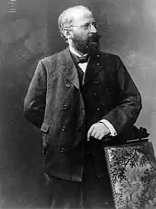 Eduard Bernstein standing next to a chair and looking rightward. He is resting his hand on the chair.