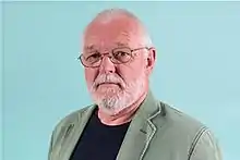 Picture of a Caucasian bearded man with gray hair and glasses standing against a blue background.