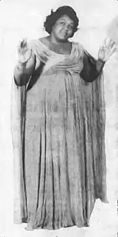 Black and white photo of an African American woman with her hands raised, who is dressed in a long flowing gown with a scooped neck.