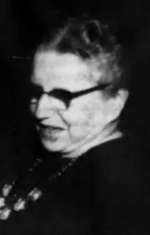 An older white woman wearing glasses, a dark top, and a necklace; her hair is center-parted and dressed back to the nape