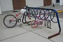 An open A-frame of thick steel tubing. Attached underneath the crossbar is a spiral of thinner metal tubing. It is painted blue, but much of the paint has flaked off, leaving rust. A mountain bike and a child's bike are parked in it and two abandoned D-locks hang from it. In the background is a brick-and-plaster building and behind the rack, to the right, is a car parking space.