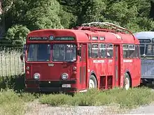 A red Wellington trolley bus with the trolleys tucked away parked up in a paddock by a fence. Bus sign says 'Karori Park number 12'.