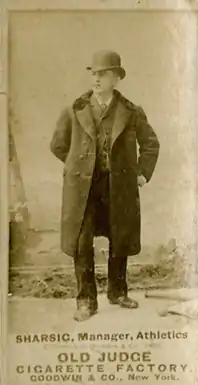 In this photograph, a man is standing facing to the right of the camera, and is wearing a suit, jacket, and a hat.