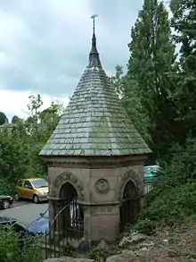 A square sandstone structure with an arch on each side, closed by a railing.  On its top is a slate spire.