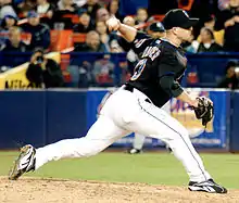 A left-handed man in his mid-thirties wearing a black baseball jersey and cap and white baseball pants throws a baseball from a pitcher's mound.