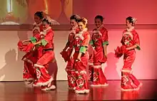 Six women in red gowns with white trim on a wooden floor against a white background performing a dance