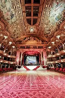 A large room with red floor and red and gold decoration to the walls.  There is a stage at the back of the picture with seating areas to the sides.