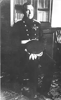 A man seated, wearing a dark uniform with two rows of buttons, with his peaked cap on his lap