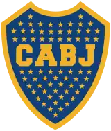 A Blue shield with a golden border. Inside the shield, 67 stars inside the shield with the golden letters "CABJ" (meaning "Club Atlético Boca Juniors") printed around the center, separating the stars