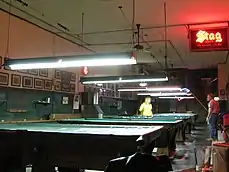 A long view of four of the pool tables at booches, some covered, and one with two players having a game; fluorescent lights and scoring strings hang above the tables.