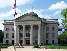 A light-colored three-story stone building with an American flag flying from a pole in front above another red-, white- and blue-striped flag. The building has a pedimented front pavilion with four columns and a small dome at the top from which another American flag is being flown.