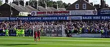 One of the stands of the Bootham Crescent association football ground, with supporters cheering and players standing on a grass field below