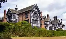 The exterior of a large house. There are several chimneys, leaded windows and wings. In the foreground are two rows of hedges.