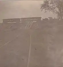 A small steam locomotive and tender, along with two passenger cars, parked in front of an open field. Stairs connect the field to the railroad tracks, which are at a higher elevation.