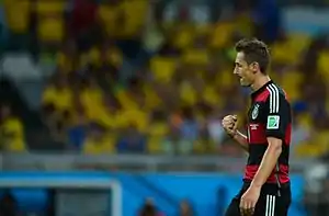 Klose pulling his arm towards himself in a goal celebration. He is wearing a red and black hooped kit. Fans in yellow Brazil jerseys are sat in the background.