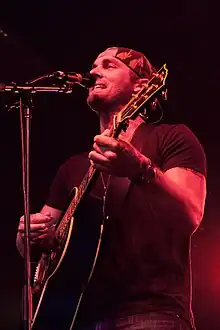 A young man wearing a dark T-shirt and cap, playing a guitar and singing into a microphone