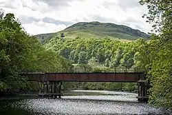 A rusty railway bridge over a dark river, with a steep hill in the distance